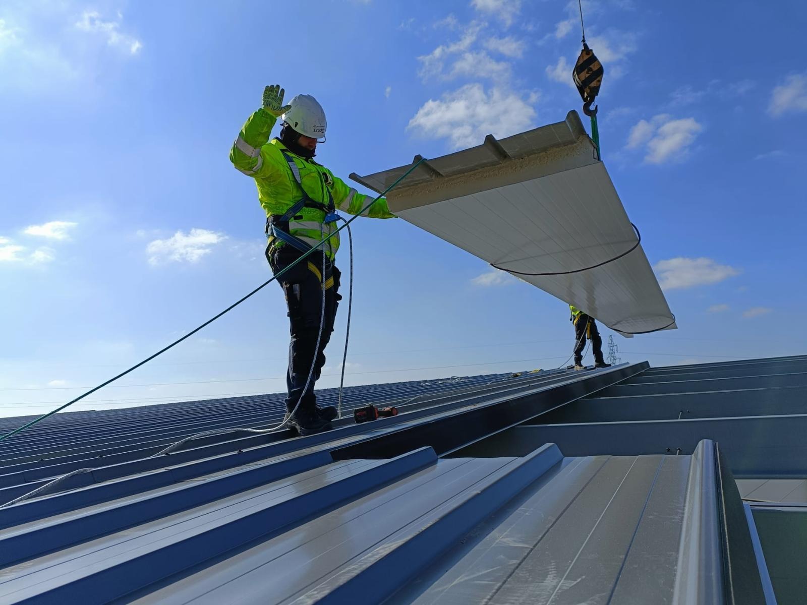Pracownik w kasku i kamizelce odblaskowej, zabezpieczony liną, montuje płytę warstwową na dachu hali; widoczny dźwig z hakiem i fragment drugiego pracownika.