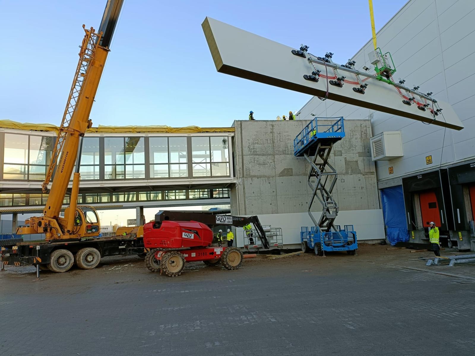 Żuraw podnosi dużą płytę warstwową za pomocą specjalistycznego chwytaka próżniowego podczas budowy hali. Widoczni pracownicy w kaskach i kamizelkach odblaskowych, podnośnik nożycowy i fragment...