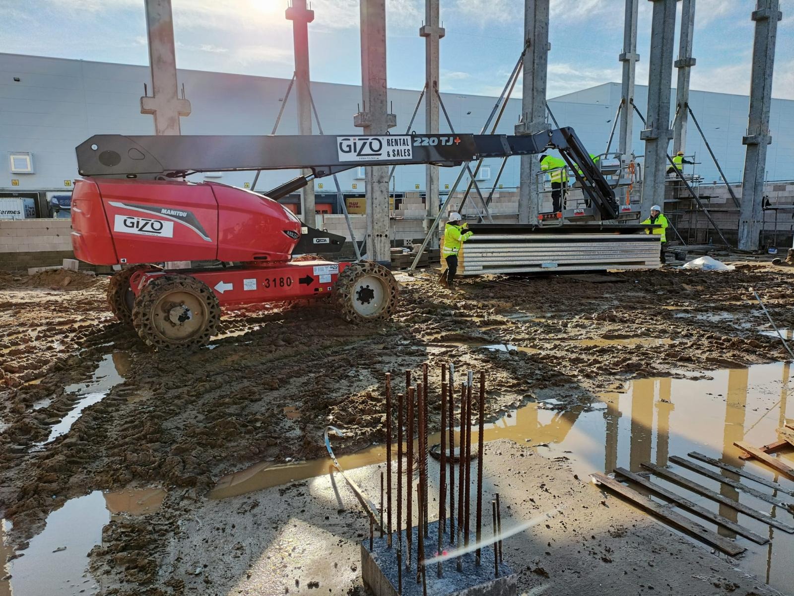 Montaż płyt warstwowych na konstrukcji hali, widoczny podnośnik GIZO, pracownicy w żółtych kamizelkach, błotnisty teren budowy.
