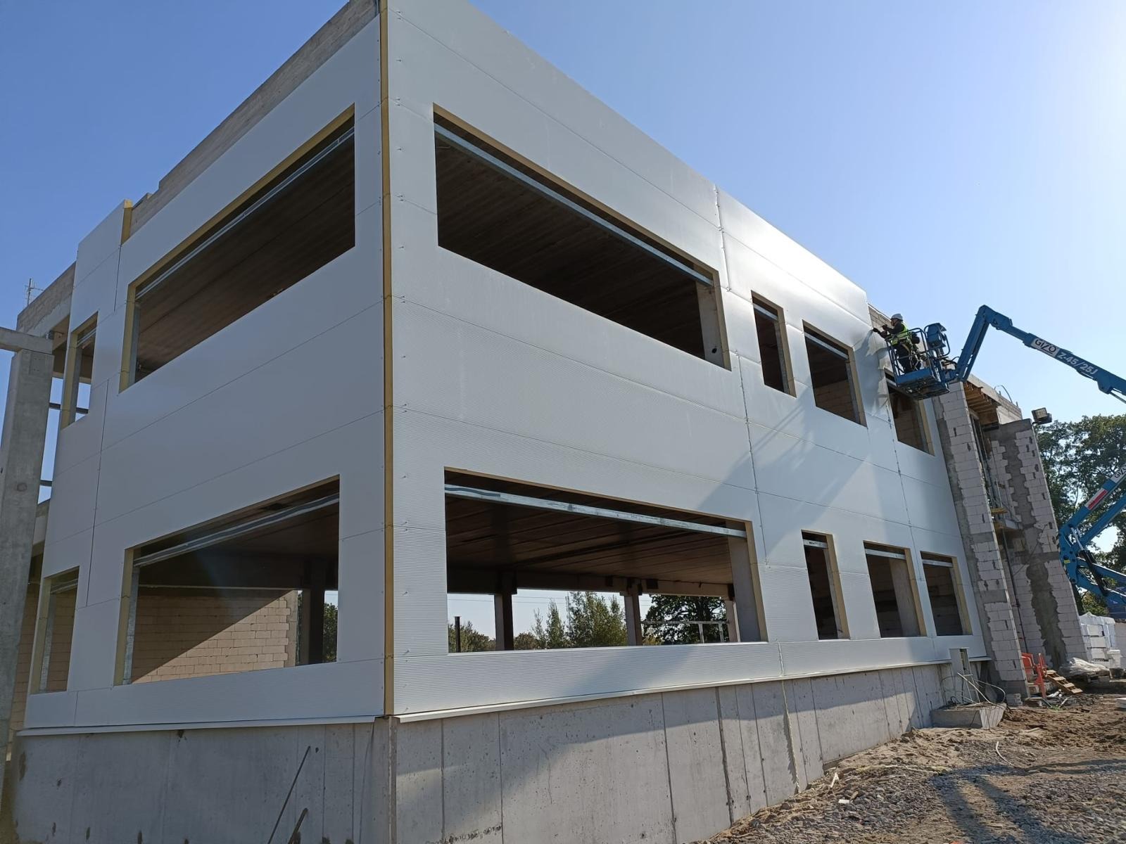 Wznoszenie dwukondygnacyjnej hali z białych płyt warstwowych, widoczny pracownik na podnośniku koszowym montujący elementy elewacji, surowy betonowy fundament i niezabudowane otwory okienne.