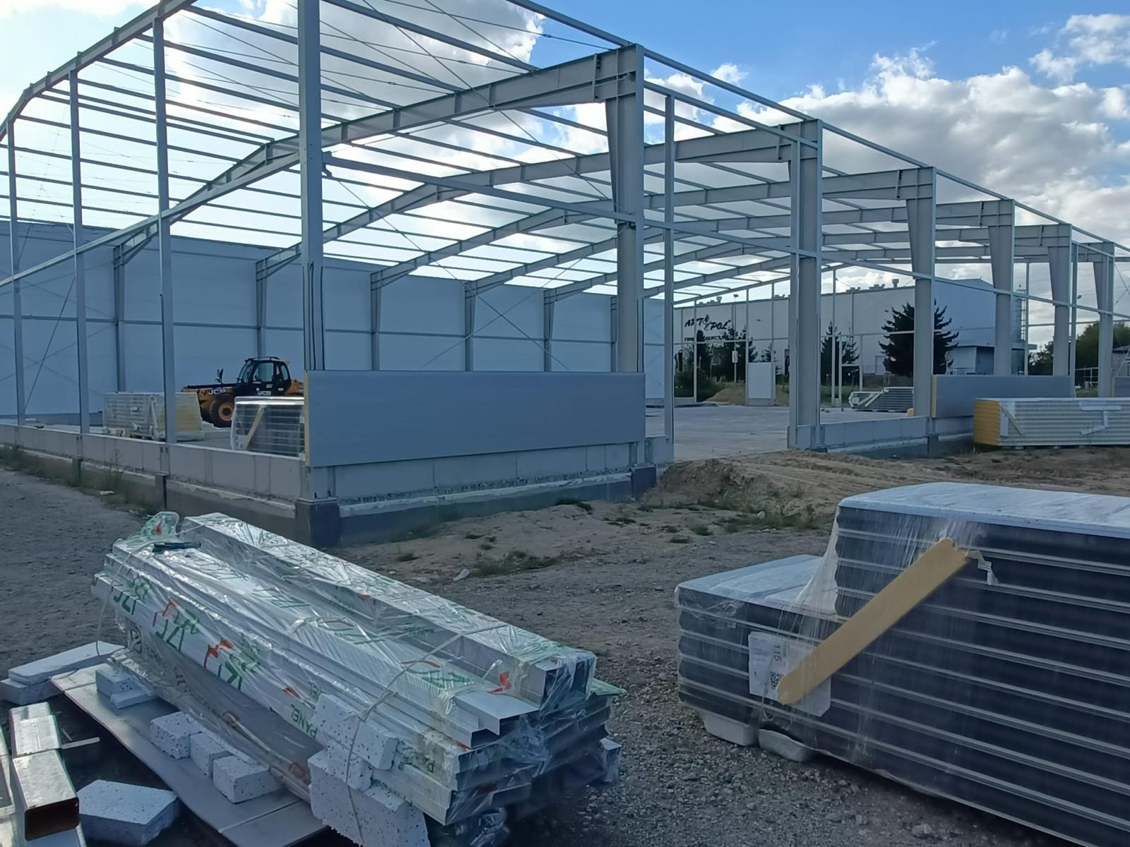 Konstrukcja stalowa hali w trakcie budowy, widoczne elementy konstrukcyjne, płyty warstwowe w folii, oraz wózek widłowy w tle pod błękitnym niebem.