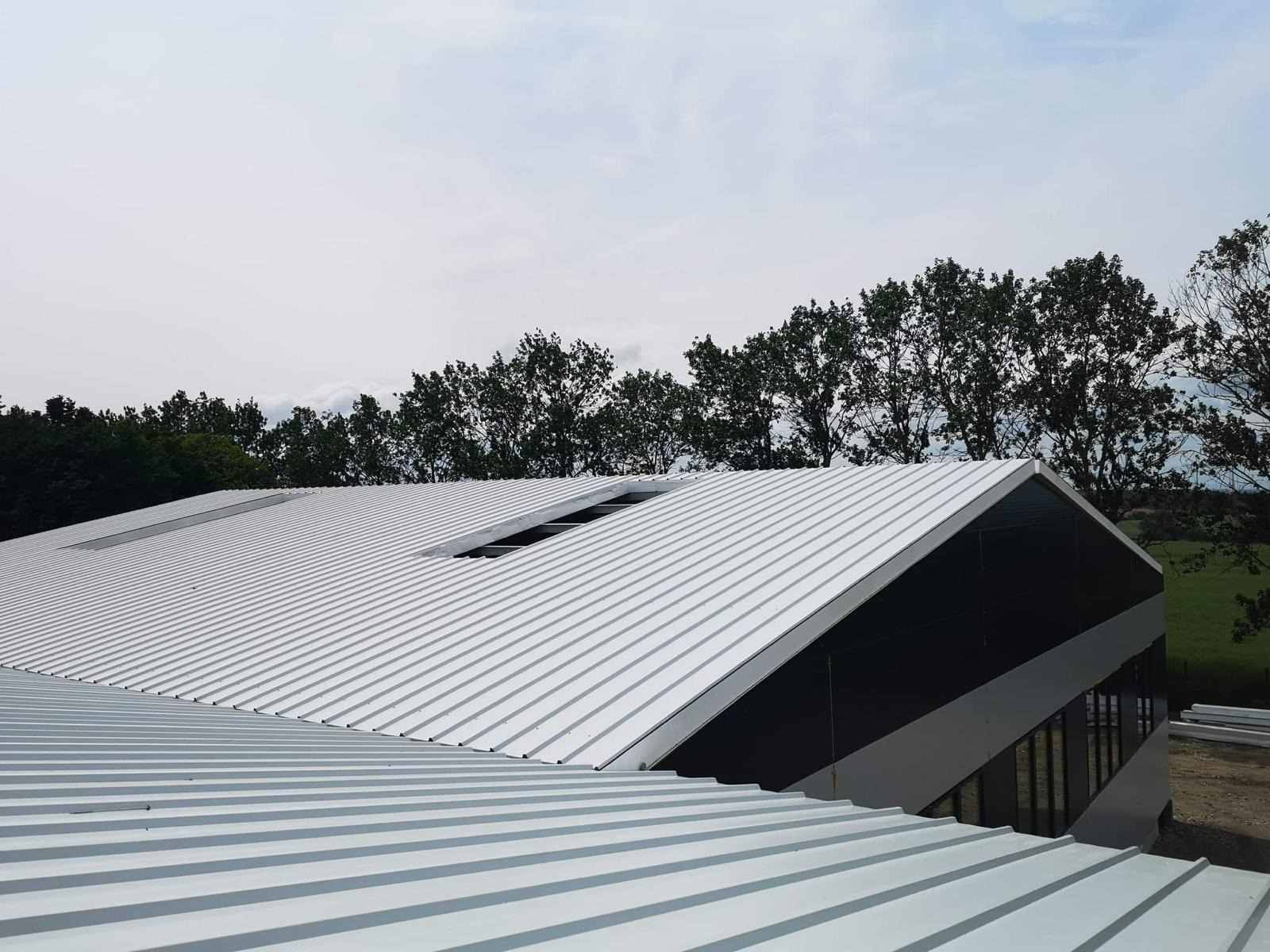 Widok z góry na metalowe poszycie dachu hali przemysłowej z wyraźnymi panelami i świetlikami, na tle zieleni drzew.