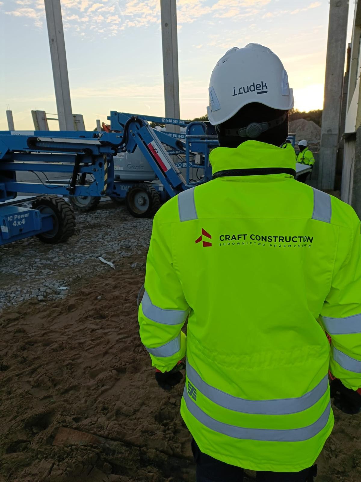 Pracownik budowlany w jaskrawej kurtce z logo Craft Construction, stojący na placu budowy hali stalowej z podnośnikami Genie w tle, betonowe słupy konstrukcyjne w trakcie montażu.