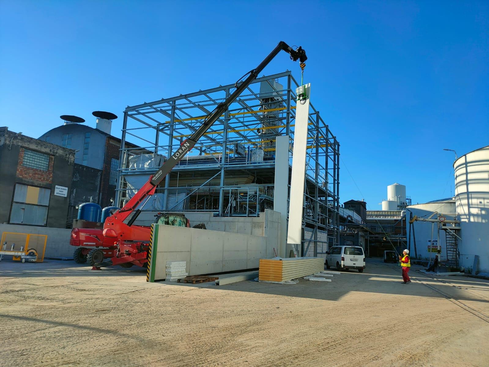 Montaż płyty warstwowej na stalowej konstrukcji hali przemysłowej z użyciem dźwigu teleskopowego, pracownik w odblaskowej kamizelce kontroluje proces, w tle widoczne zbiorniki i budynki przemysłowe...