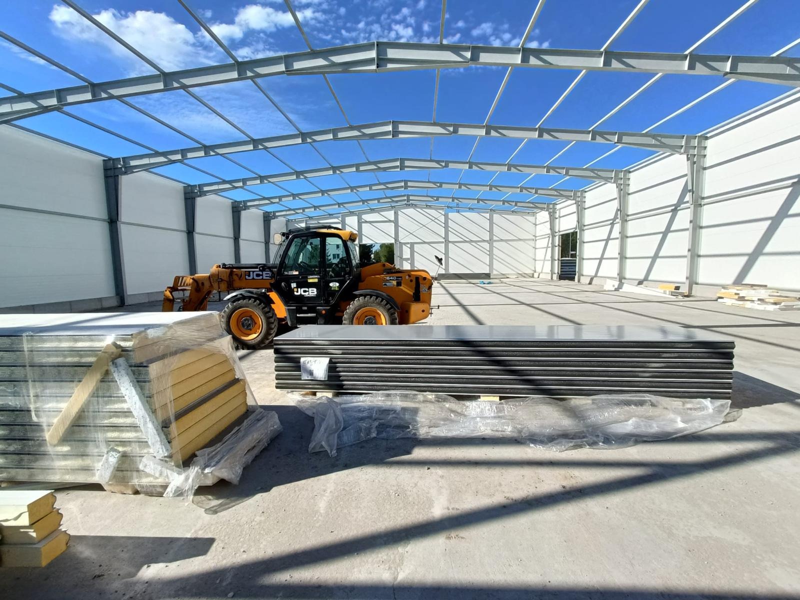 Wnętrze wznoszonej hali z widoczną stalową konstrukcją, stertą płyt warstwowych w folii i pomarańczową ładowarką teleskopową JCB na betonowej posadzce w słoneczny dzień.