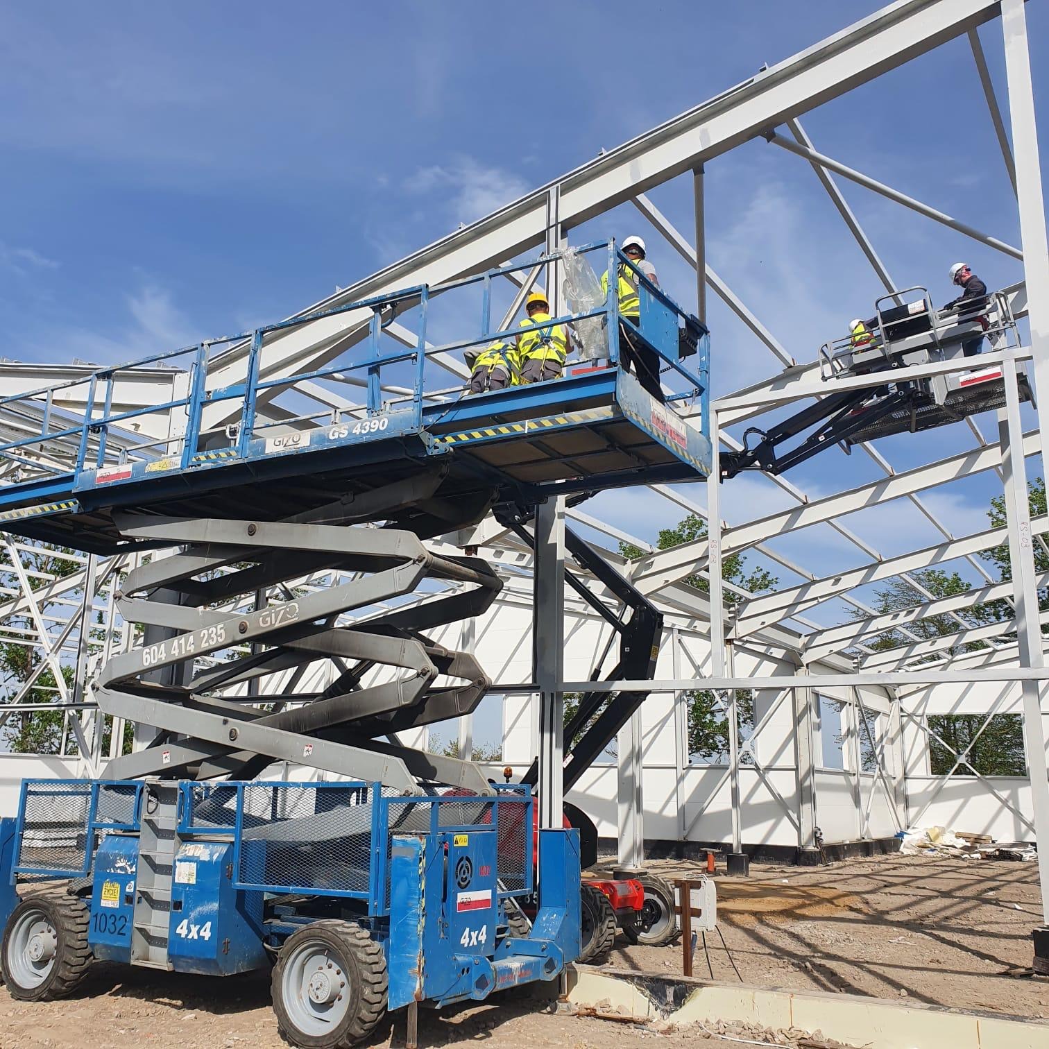 Montaż konstrukcji stalowej hali w Szczecinie z wykorzystaniem podnośników nożycowych i koszowych; widoczni pracownicy w kaskach i kamizelkach odblaskowych na tle błękitnego nieba.