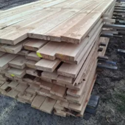 Stos desek z jasnego drewna ułożonych na przekładkach, widoczne żółte oznaczenia na niektórych deskach, w tle plac z maszynami budowlanymi.