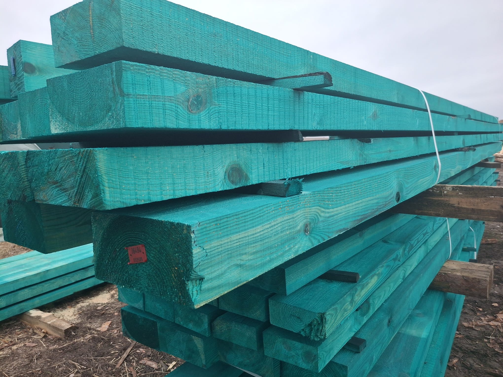 Stos impregnowanych na turkusowo kantówek z widocznymi numerami partii na czerwonych etykietach, ułożonych na drewnianych legarach na zewnątrz.