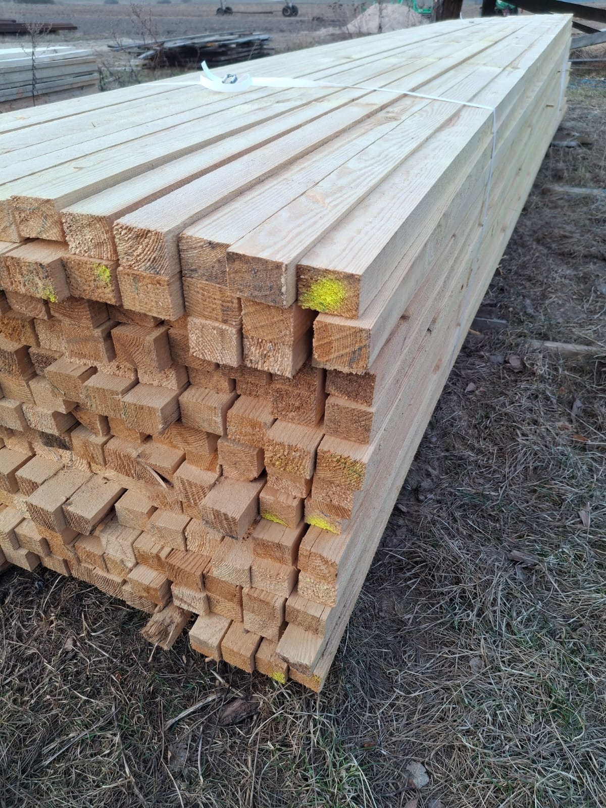 Stos świeżo przetartego drewna budowlanego, zabezpieczonego taśmą, z widocznymi żółtymi oznaczeniami na końcach, leżący na wysuszonej trawie.