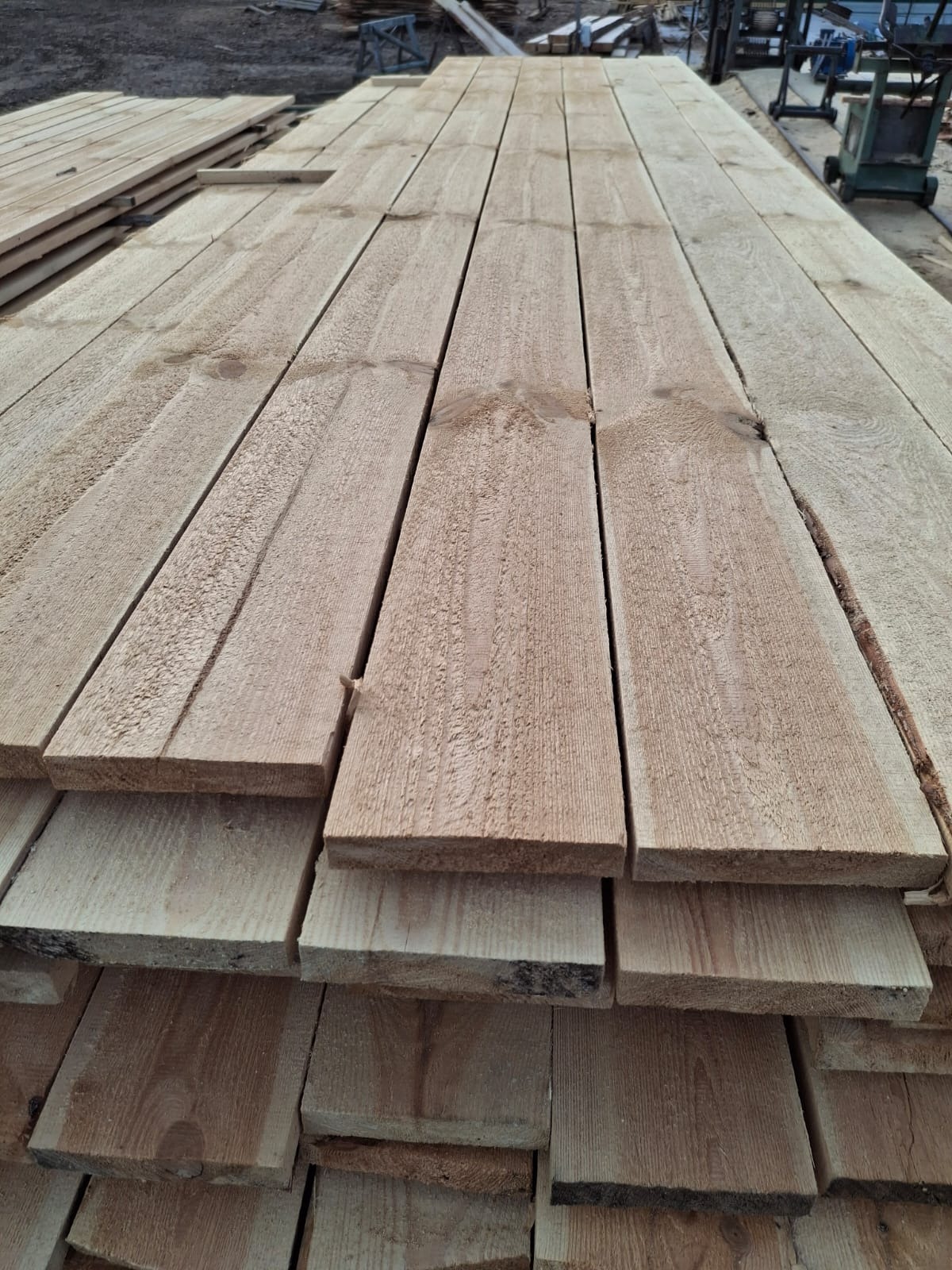 Stos desek z surowego drewna ułożonych warstwami, widoczne sęki i naturalna faktura, w tle maszyna do obróbki drewna.