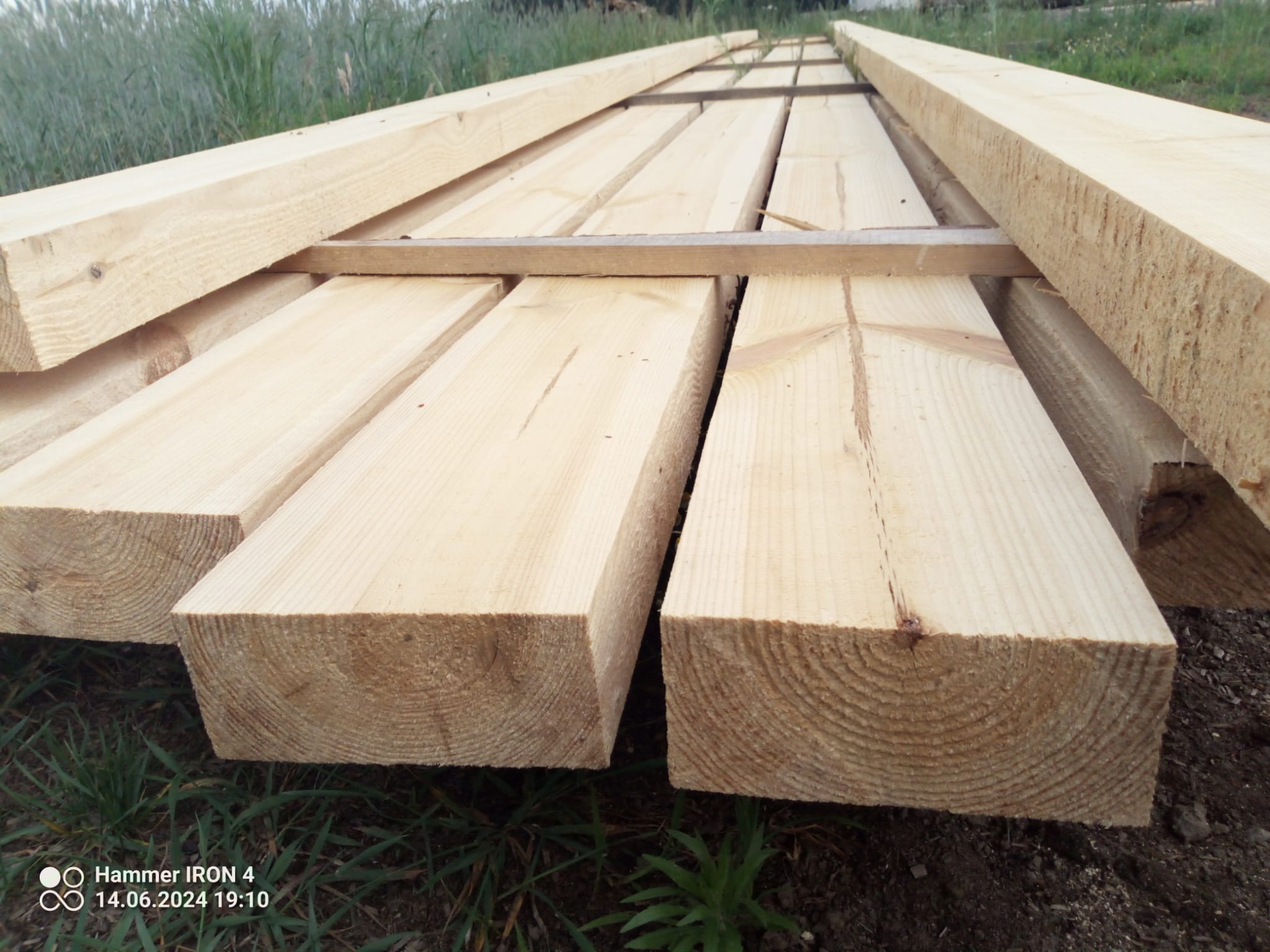 Stos świeżo przetartego, jasnego drewna budowlanego ułożonego na przekładkach, widoczny na tle trawy.