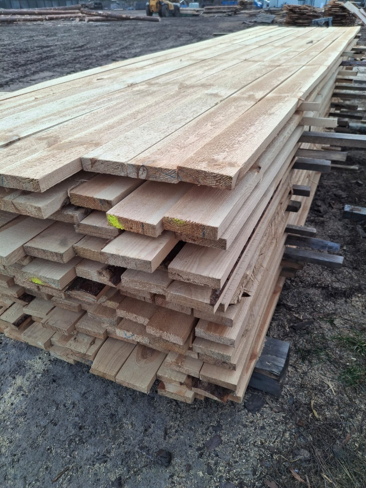 Stos desek z jasnego drewna ułożonych na przekładkach, widoczne żółte oznaczenia na niektórych deskach, w tle plac z maszynami budowlanymi.