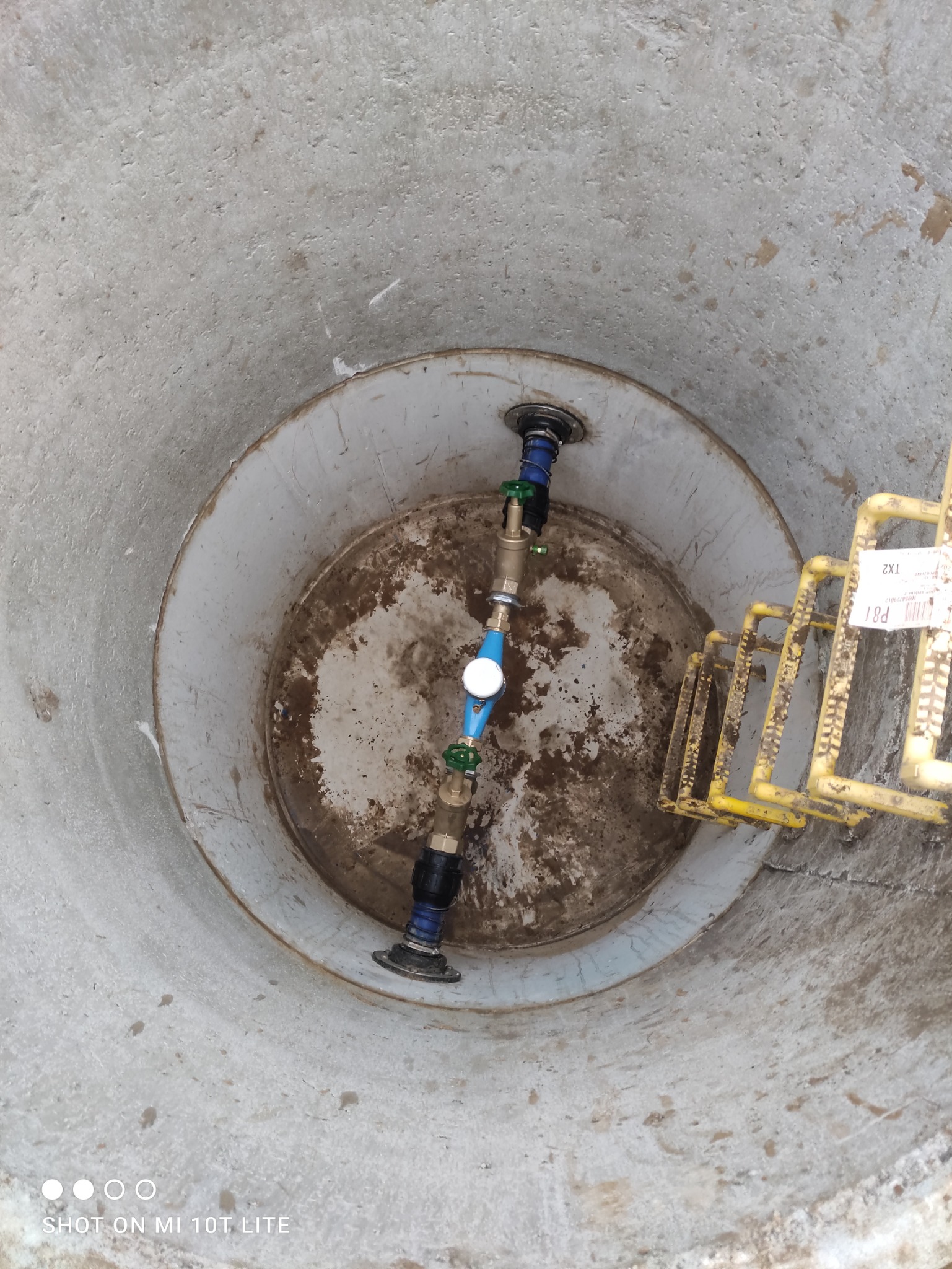 Wnętrze betonowej studni z zainstalowanym przyłączem wodnym i fragmentem drabiny zejściowej. Widok z góry, ukazujący detale instalacji i surowość betonowej konstrukcji.