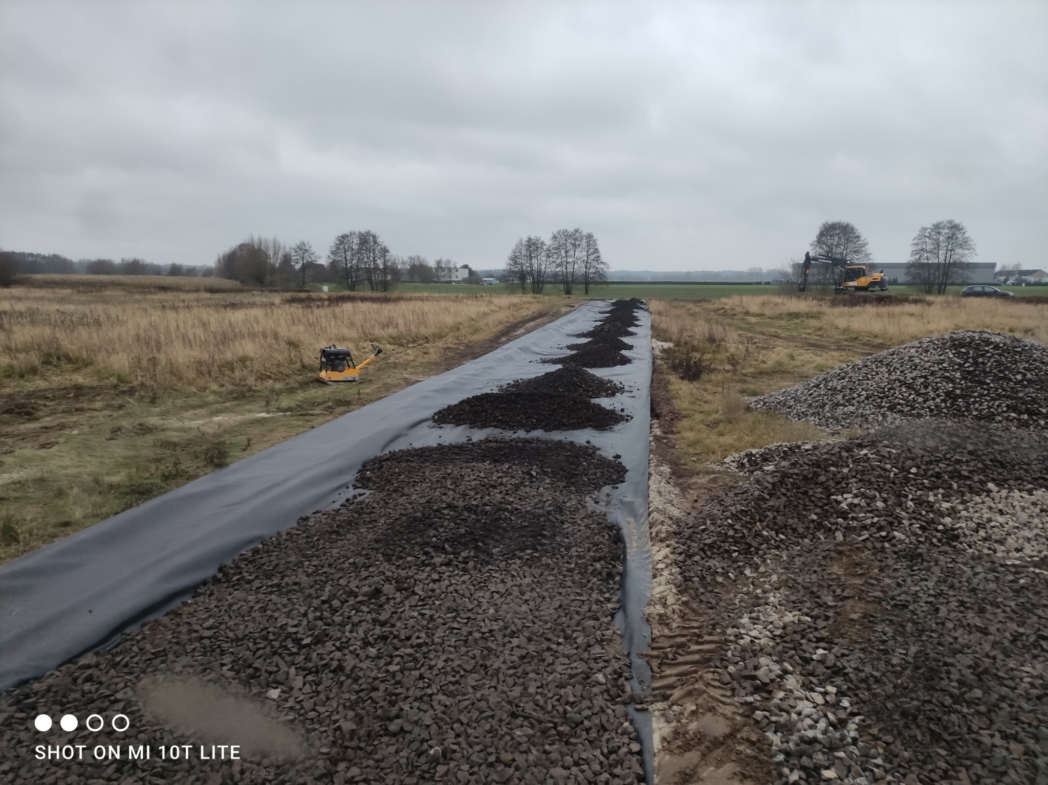 Proces utwardzania drogi: rozłożona geowłóknina, nasypy kamienia, ubijarka i koparka w tle na tle pochmurnego nieba. Widok z góry.