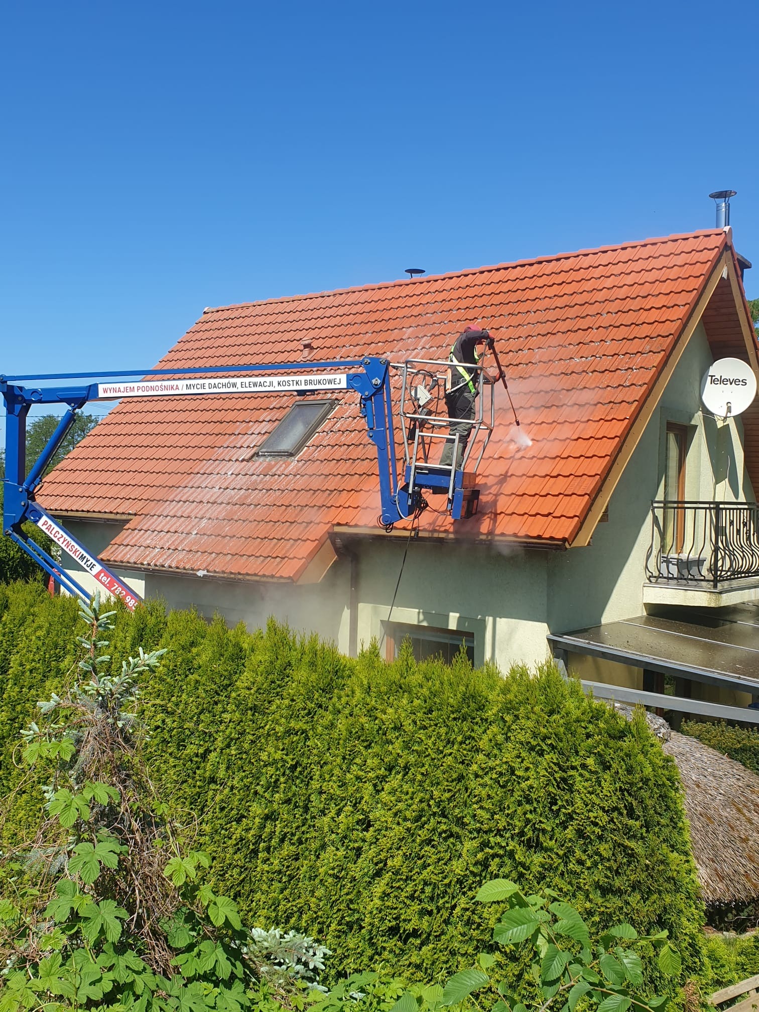 Pracownik w podnośniku koszowym czyści dach domu z czerwonej dachówki za pomocą myjki ciśnieniowej, widoczne strumienie wody i para, w tle błękitne niebo i zielone tuje.