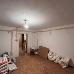 Sebastian Łęcki - Usługi elektryczne - Pokój w trakcie remontu, z widocznymi śladami po instalacji elektrycznej na ścianach i suficie, wiszącymi przewodami, workami z zaprawą gipsową i prowizorycznym oświetleniem w postaci żarówki...
