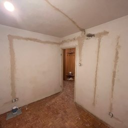 Sebastian Łęcki - Usługi elektryczne - Pokój w trakcie remontu: ściany z widocznymi śladami po instalacjach elektrycznych i świeżo położonej gładzi, odsłonięte gniazdka elektryczne, parkiet na podłodze, widoczne drzwi do innego...