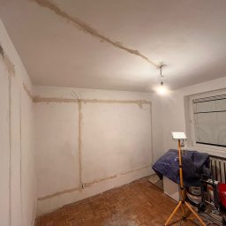 Sebastian Łęcki - Usługi elektryczne - Pokój w trakcie remontu: widoczne ślady po instalacji elektrycznej na ścianach i suficie, przygotowane do dalszych prac wykończeniowych, z lampą roboczą na statywie i materiałami budowlanymi w tle.
