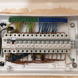 Sebastian Łęcki - Usługi elektryczne - Otwarta skrzynka bezpiecznikowa z widocznymi bezpiecznikami marki Eaton i Legrand oraz przewodami w kolorach zielonym, żółtym, niebieskim i brązowym, zamontowana na ścianie z widocznymi śladami po...