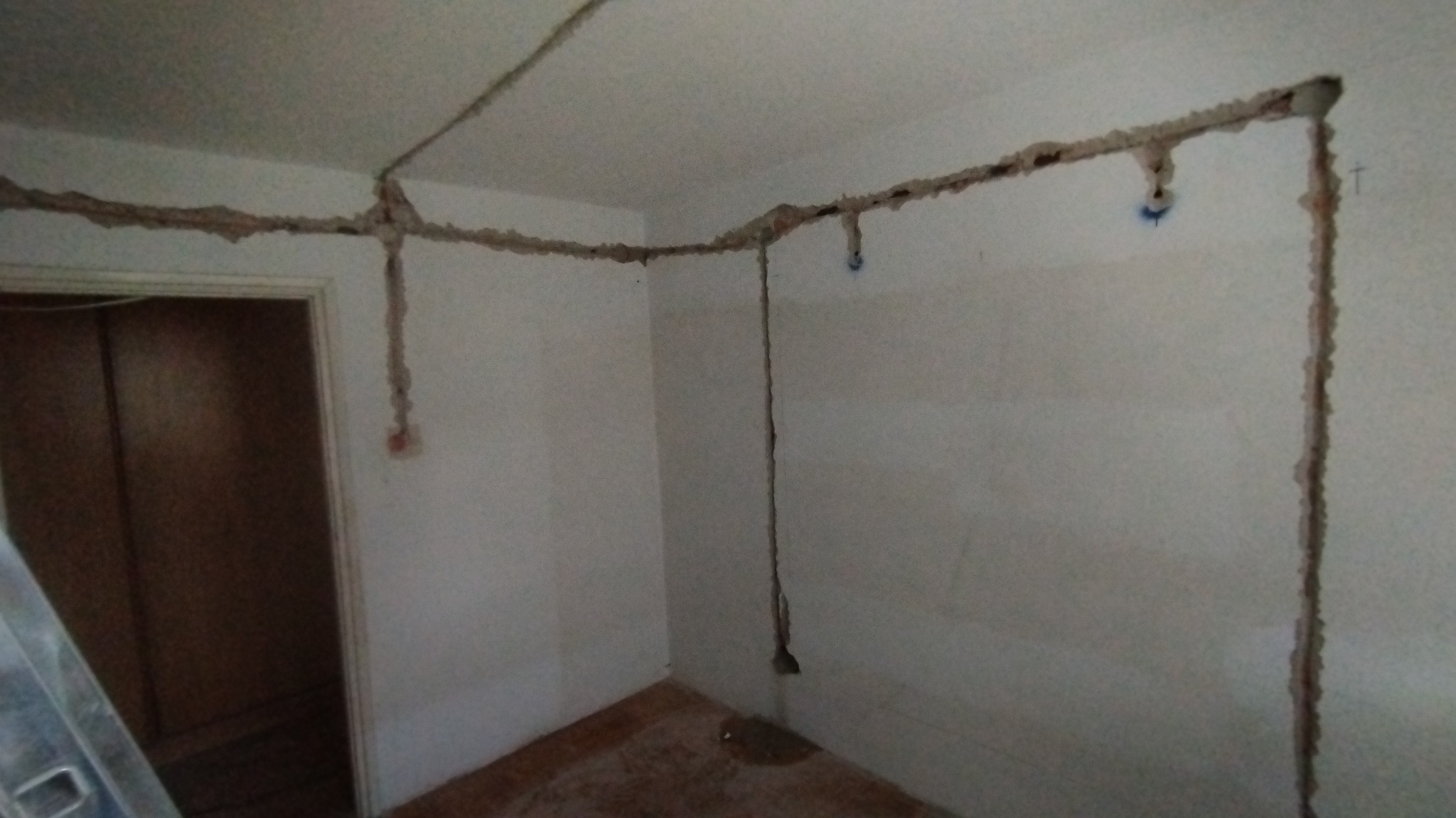 Wyżłobienia w ścianach i suficie przygotowane pod instalację elektryczną, widoczne kable wystające ze ściany, fragment drabiny malarskiej w rogu pomieszczenia.