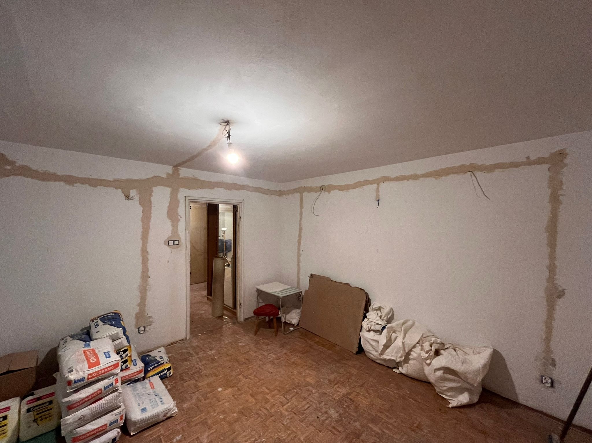 Pokój w trakcie remontu, z widocznymi śladami po instalacji elektrycznej na ścianach i suficie, wiszącymi przewodami, workami z zaprawą gipsową i prowizorycznym oświetleniem w postaci żarówki...