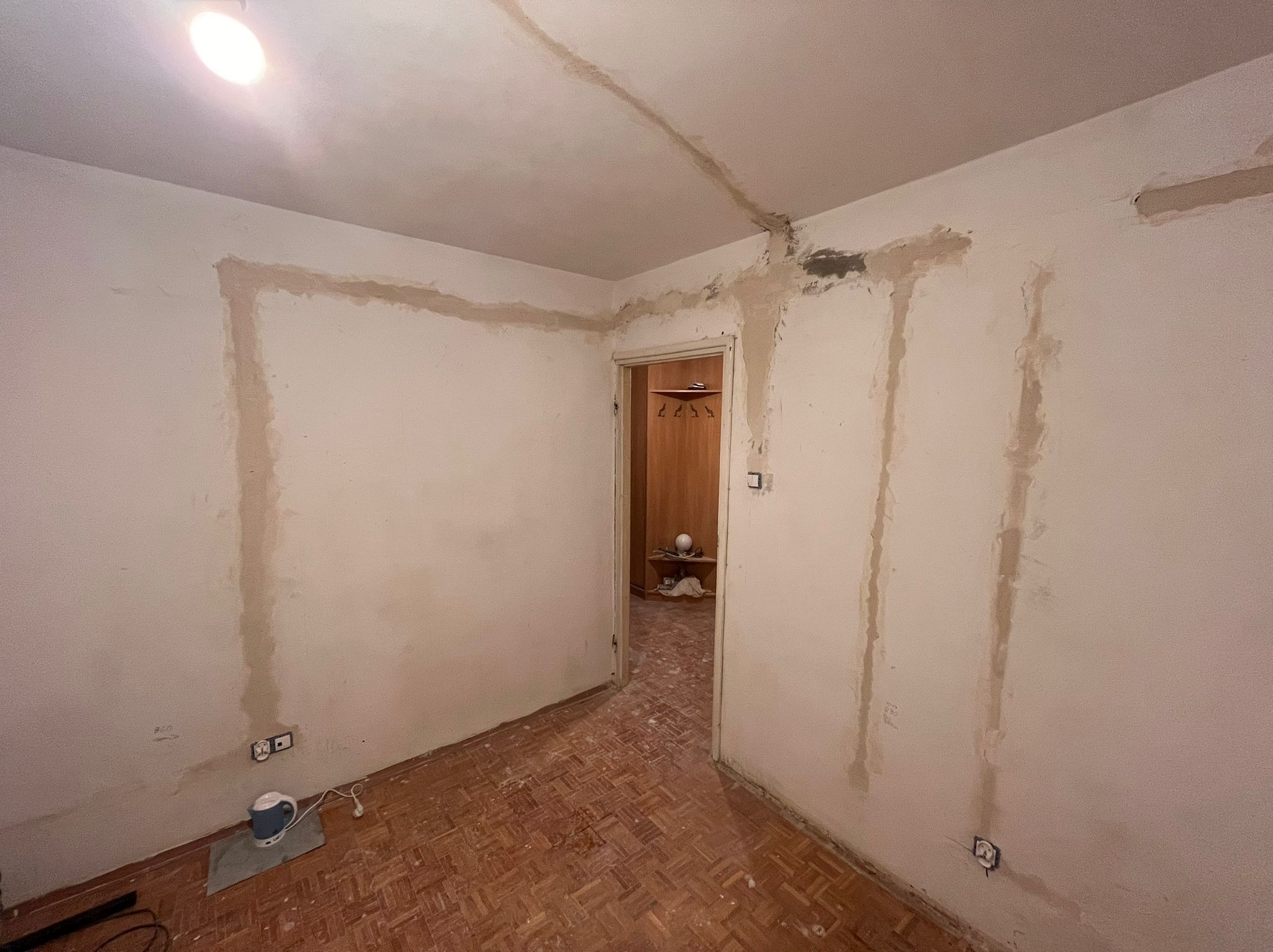 Pokój w trakcie remontu: ściany z widocznymi śladami po instalacjach elektrycznych i świeżo położonej gładzi, odsłonięte gniazdka elektryczne, parkiet na podłodze, widoczne drzwi do innego...