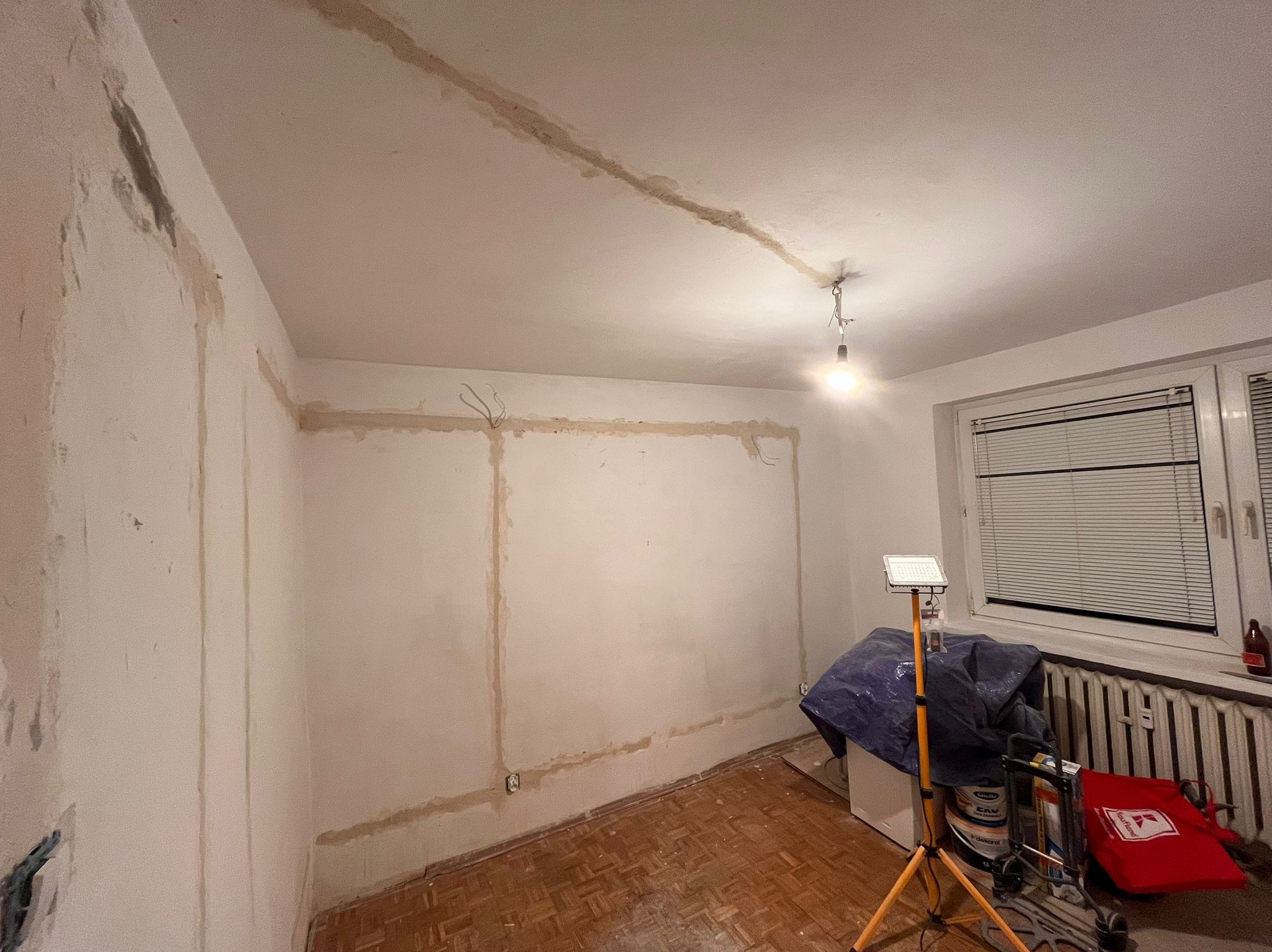 Pokój w trakcie remontu: widoczne ślady po instalacji elektrycznej na ścianach i suficie, przygotowane do dalszych prac wykończeniowych, z lampą roboczą na statywie i materiałami budowlanymi w tle.