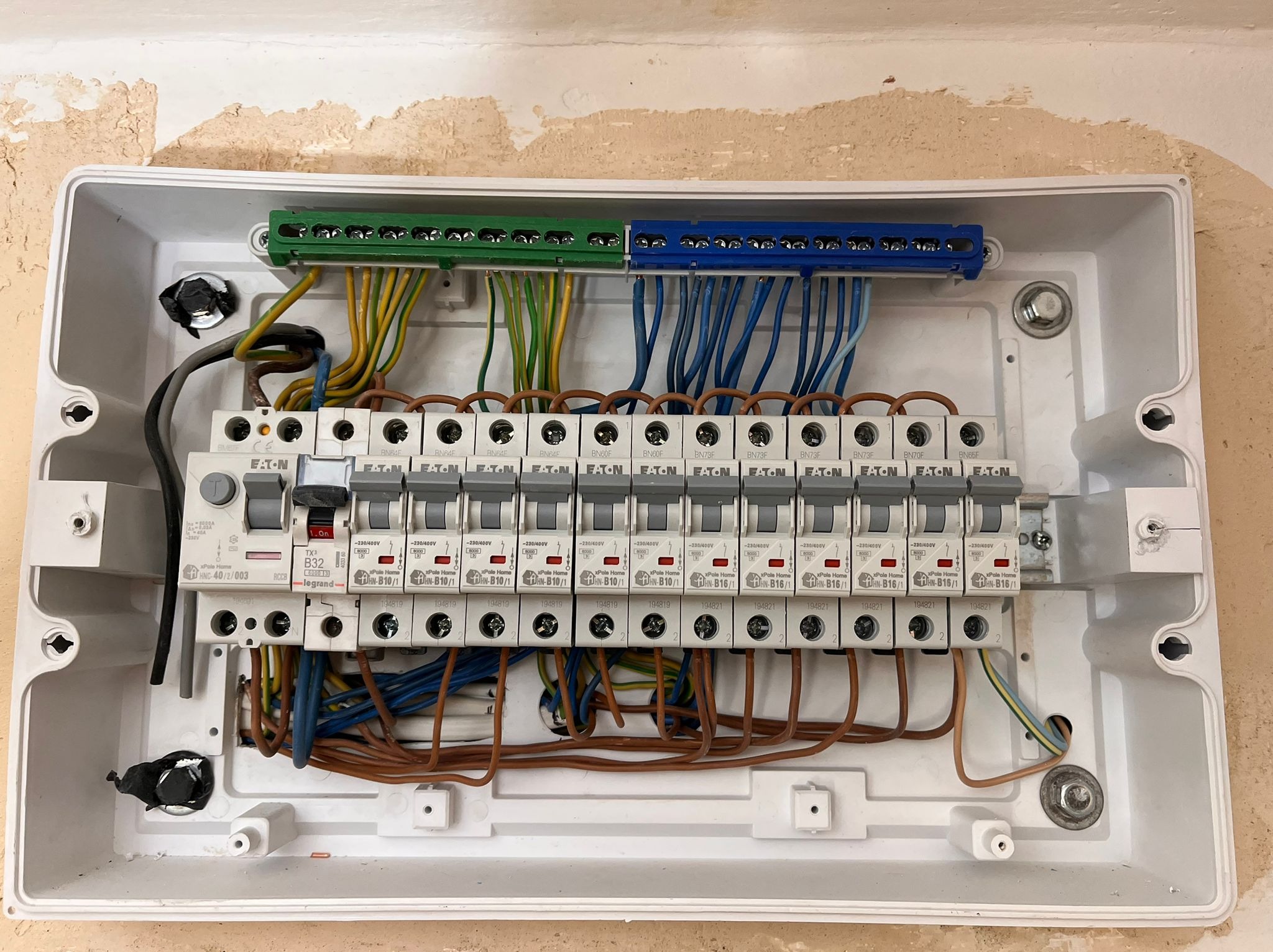Otwarta skrzynka bezpiecznikowa z widocznymi bezpiecznikami marki Eaton i Legrand oraz przewodami w kolorach zielonym, żółtym, niebieskim i brązowym, zamontowana na ścianie z widocznymi śladami po...