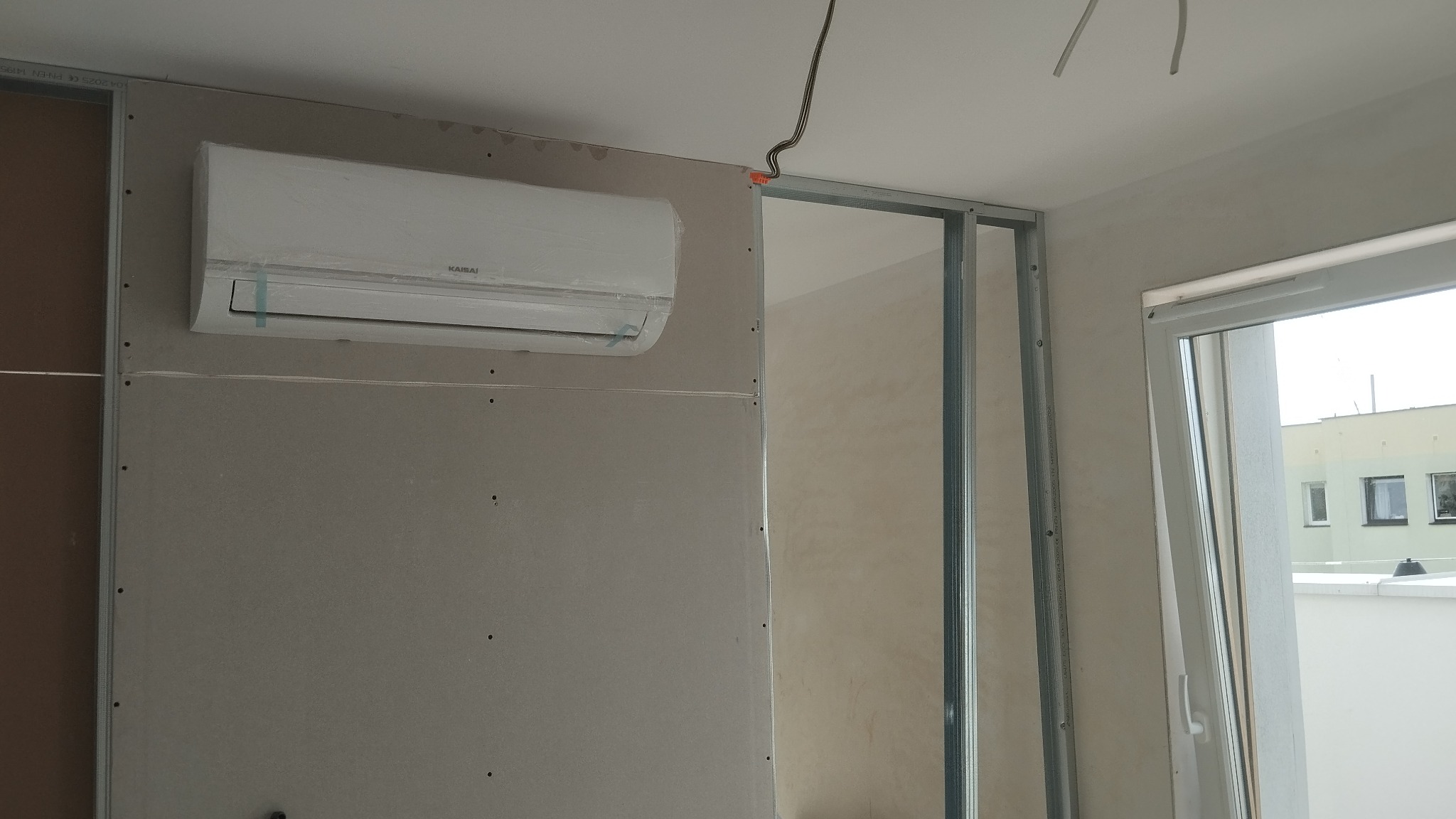 Biały klimatyzator na ścianie z płyt gipsowo-kartonowych, widoczne profile metalowe. W tle okno z widokiem na budynek. Instalacja w trakcie wykończenia.