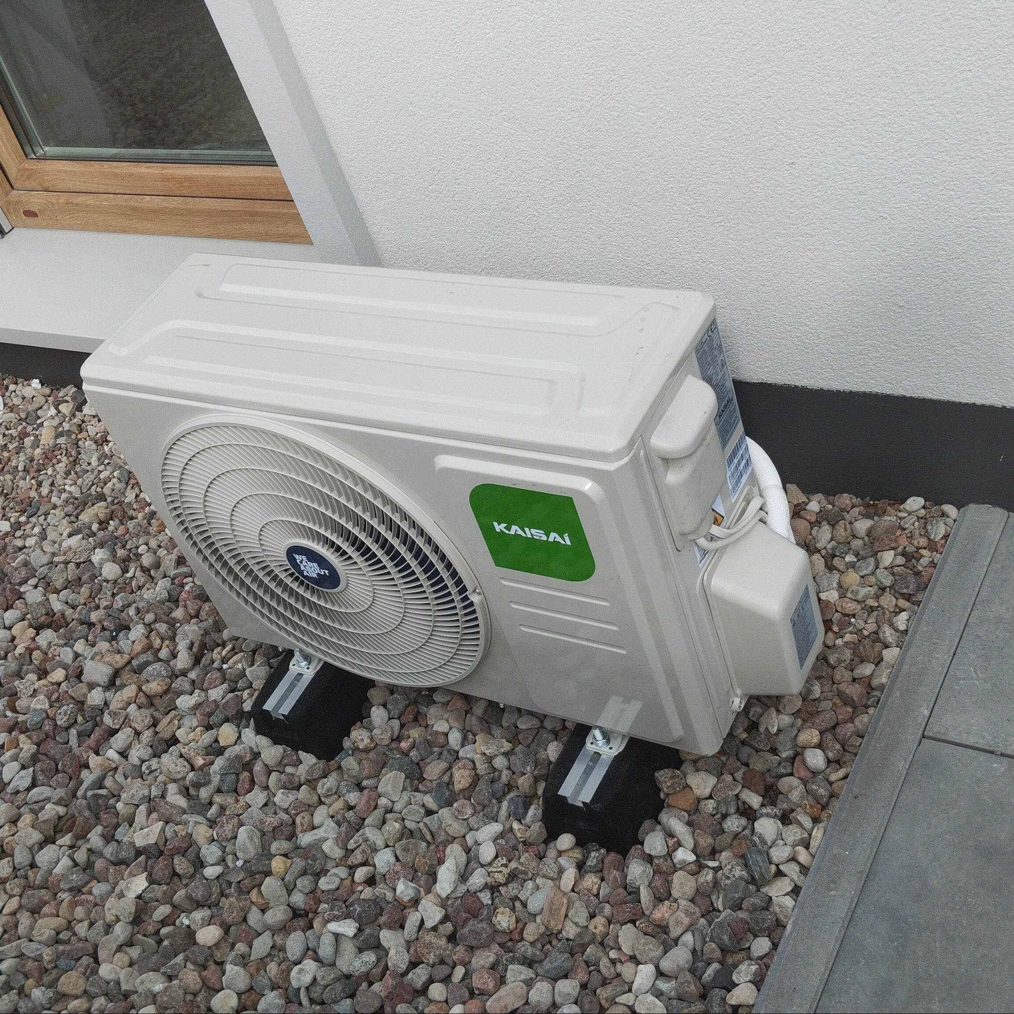 Kompaktowa, zewnętrzna jednostka klimatyzacji Kaisai, zamontowana na czarnych wspornikach, otoczona ozdobnymi kamieniami, przy ścianie budynku z oknem.