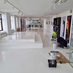 RDK. Serwis - Dwóch mężczyzn w trakcie wykonywania prac wykończeniowych w jasnym, przestronnym biurze z białymi ścianami i podłogą. Jeden mężczyzna stoi i rozprowadza masę wałkiem, drugi klęczy obok wiadra...