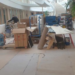 RDK. Serwis - Wnętrze budynku w trakcie prac wykończeniowych: widoczny pracownik szlifujący ścianę, podnośnik, kartony, narzędzia i materiały budowlane na podłodze, w tle spiralne schody.