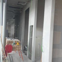RDK. Serwis - Wnętrze biurowe w trakcie prac: widoczne surowe ściany z nałożoną warstwą szpachli, odsłonięte elementy instalacji pod sufitem, materiały budowlane i narzędzia na podłodze, przewody elektryczne.