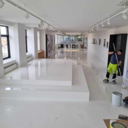 RDK. Serwis - Pracownik w jaskrawożółtych spodniach maluje jasną podłogę w przestronnym biurze z oknami i reflektorami sufitowymi, widoczny kubeł z farbą i wałek.