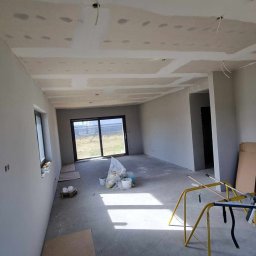 RDK. Serwis - Wnętrze w trakcie remontu: surowy stan z widocznymi gładziami na suficie, worki z materiałami budowlanymi, żółty kozioł malarski i częściowo zabezpieczona podłoga kartonem.