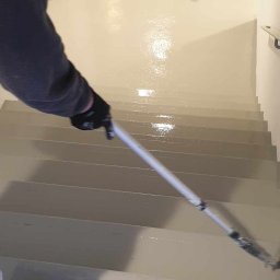 RDK. Serwis - Malowanie schodów wałkiem teleskopowym na jasnoszaro, widoczne świeże ślady farby i robocze obuwie malarza.