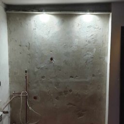 RDK. Serwis - Ściana w trakcie przygotowywania pod dalsze prace wykończeniowe, widoczne nierówności i ślady po wcześniejszych instalacjach, oświetlenie punktowe podkreśla fakturę powierzchni.