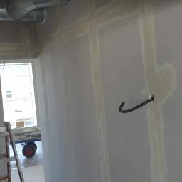 RDK. Serwis - Ściana z płyt gipsowo-kartonowych, z widocznymi łączeniami zabezpieczonymi taśmą i wypełnionymi masą szpachlową, w tle drabina malarska i wózek transportowy.