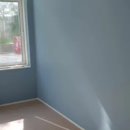 Jasne, błękitne ściany w pokoju biurowym z wykładziną dywanową, widoczne okno z białymi ramami i fragment sufitu podwieszanego z otwartym panelem.