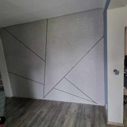 RDK. Serwis - Ściana z gładzią w Chełmnie z geometrycznym wzorem z czarnych linii, widoczny fragment pokoju z drzwiami i salon z sofą w tle.