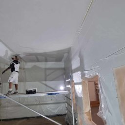 RDK. Serwis - Pracownik na rusztowaniu wyrównuje sufit w pomieszczeniu zabezpieczonym folią malarską, widoczny fragment ściany z oknem.