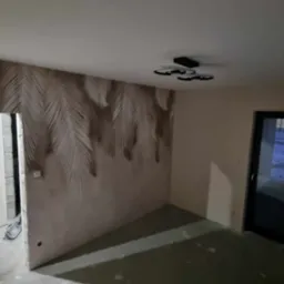 Pokój po tapetowaniu z motywem palmowych liści na jednej ścianie, widoczna nowoczesna lampa sufitowa i drzwi balkonowe, w tle widok na zaśnieżony krajobraz.