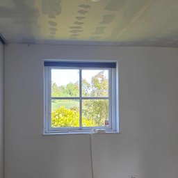 RDK. Serwis - Pokój przed remontem: widok na sufit z widocznymi śladami po szpachlowaniu, jasne ściany, drewniana podłoga, okno z widokiem na zieleń.