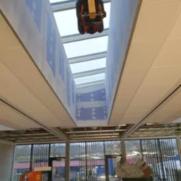 Wnętrze w trakcie prac: widok na sufit z oknem dachowym, częściowo pokryty płytami gipsowymi, z widocznymi śladami szpachlowania i szlifierką do gipsu zawieszoną pod oknem dachowym. Na dole...