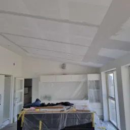 Wnętrze w trakcie remontu: sufit z perforowanymi panelami częściowo pokryty gładzią, meble zabezpieczone folią malarską, widoczne narzędzia i materiały przygotowane do dalszych prac.