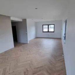 Jasne, przestronne wnętrze po remoncie z podłogą ułożoną w jodełkę i świeżo pomalowanymi ścianami na biało. Okno z czarną ramą i widokiem na zewnątrz.