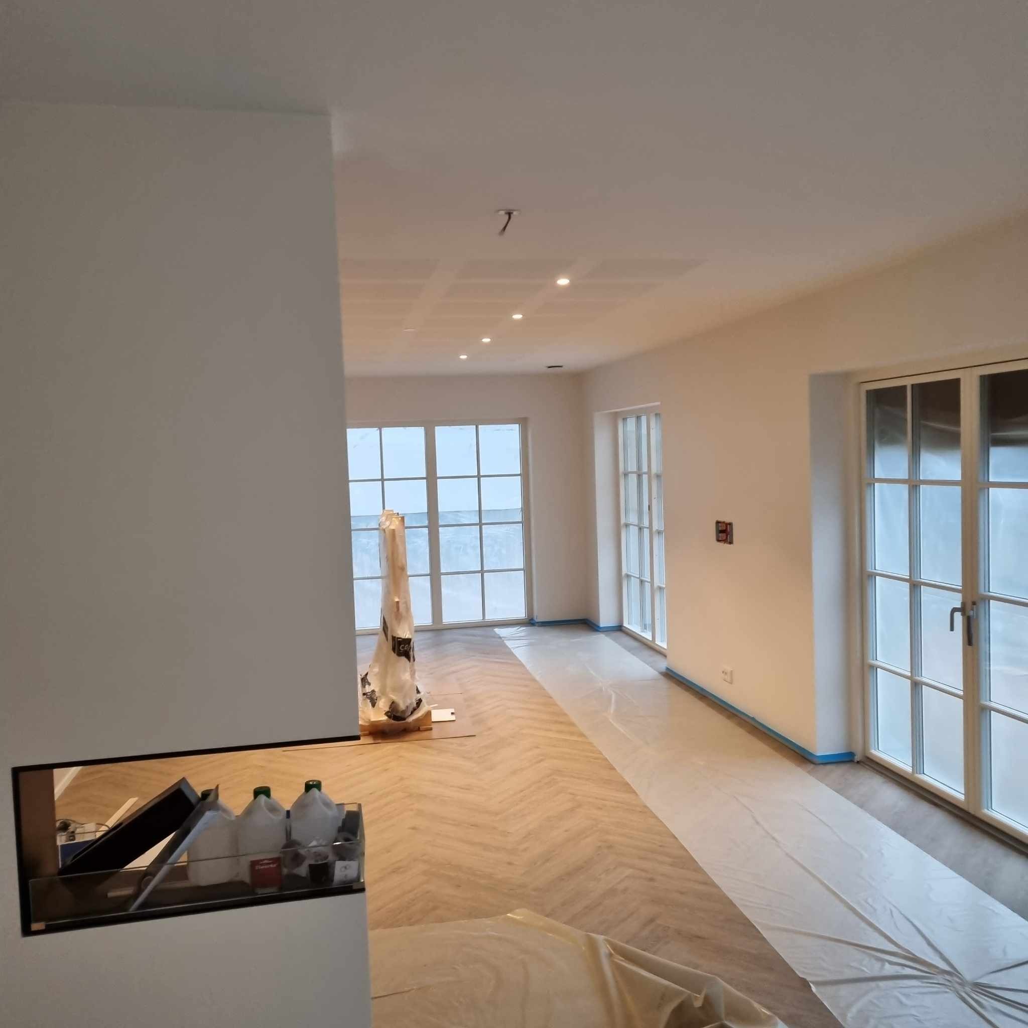 Przygotowane pomieszczenie do prac malarskich, podłoga zabezpieczona folią, ściany świeżo pomalowane na biało, widoczne okna z niebieską taśmą malarską.