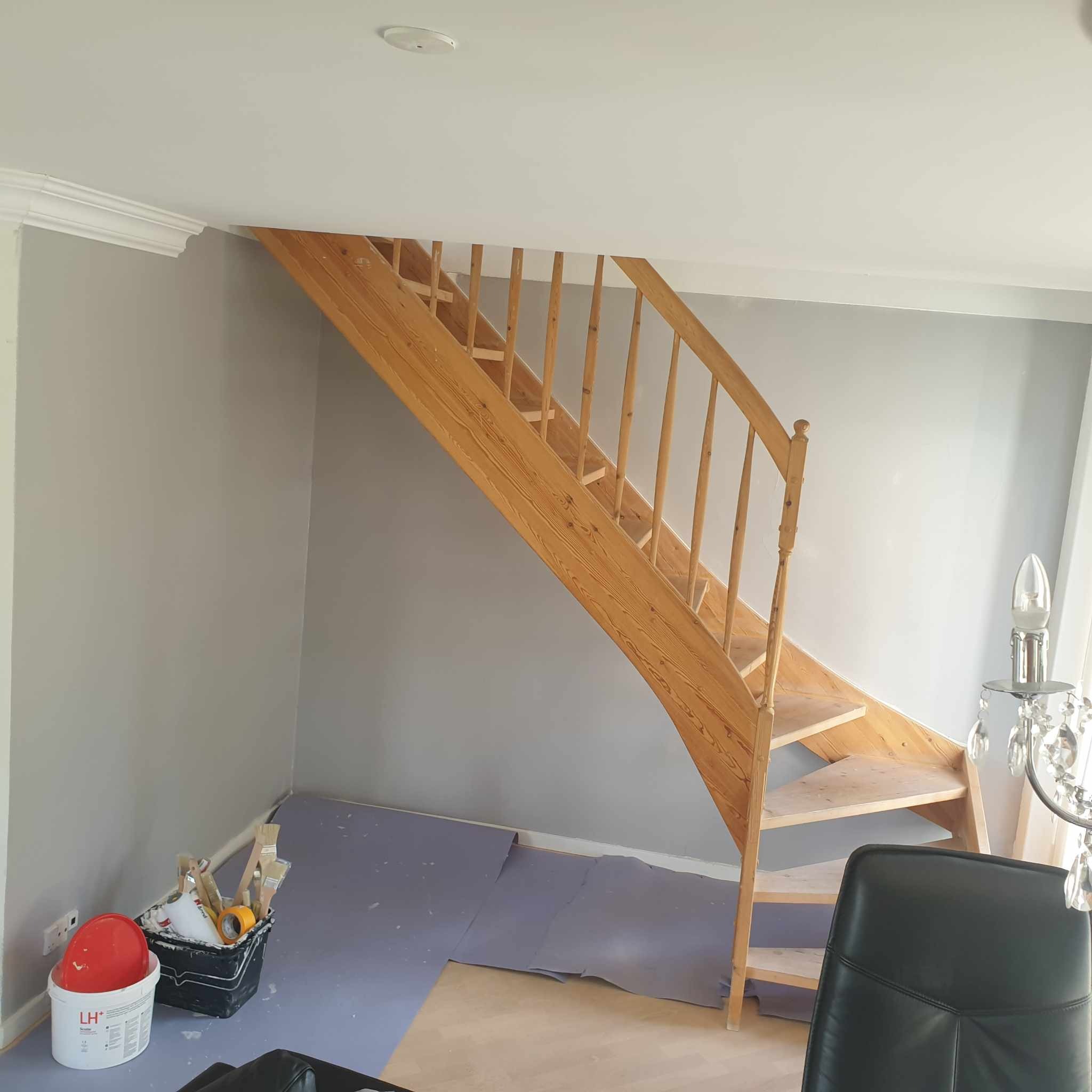 Drewniane schody w trakcie renowacji, zabezpieczona podłoga, ściany pomalowane na szaro, widoczne narzędzia malarskie w wiadrze.