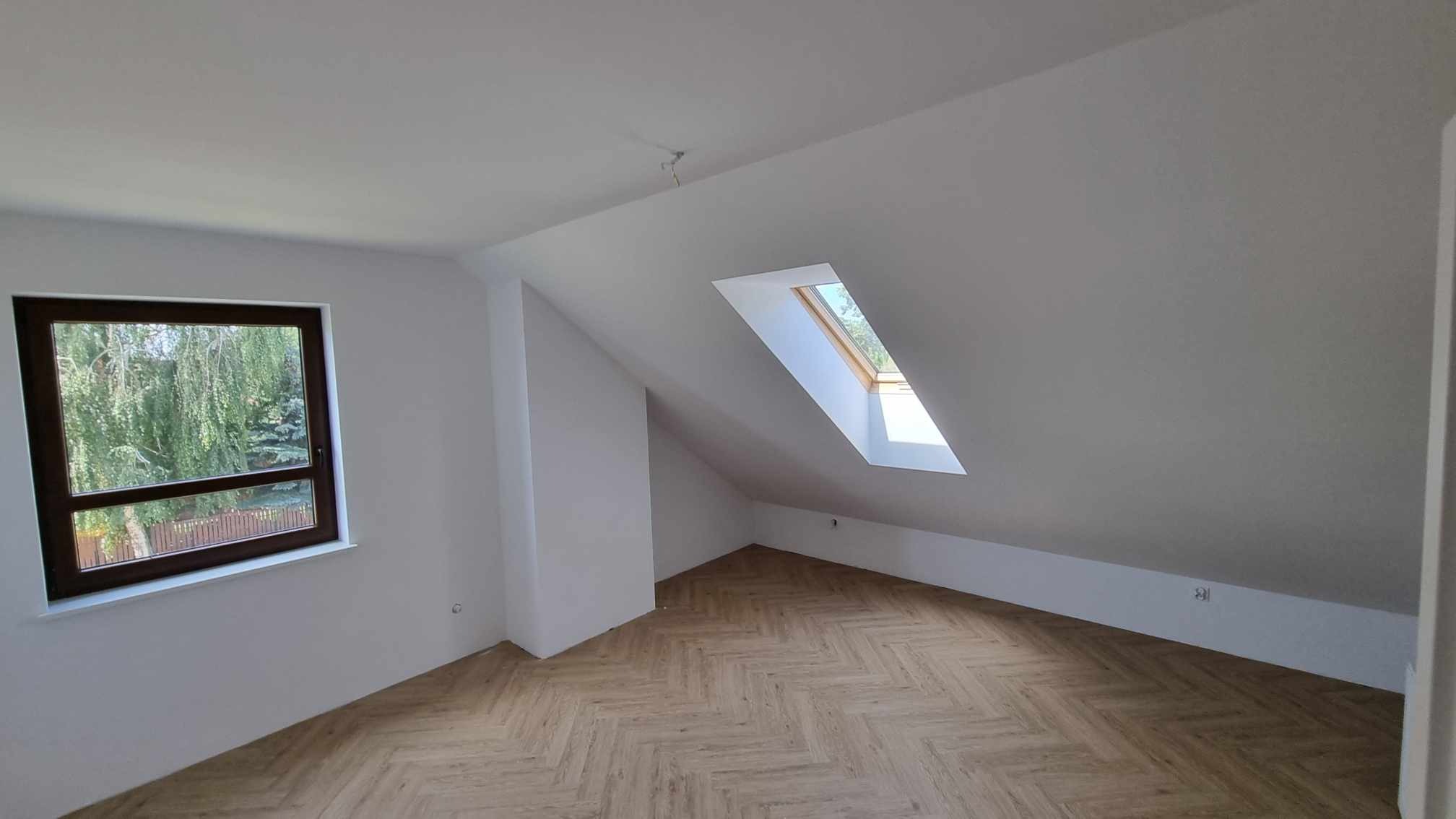 Poddasze po remoncie z jasnymi ścianami, drewnianą podłogą ułożoną w jodełkę i oknem dachowym oraz oknem na ścianie szczytowej.