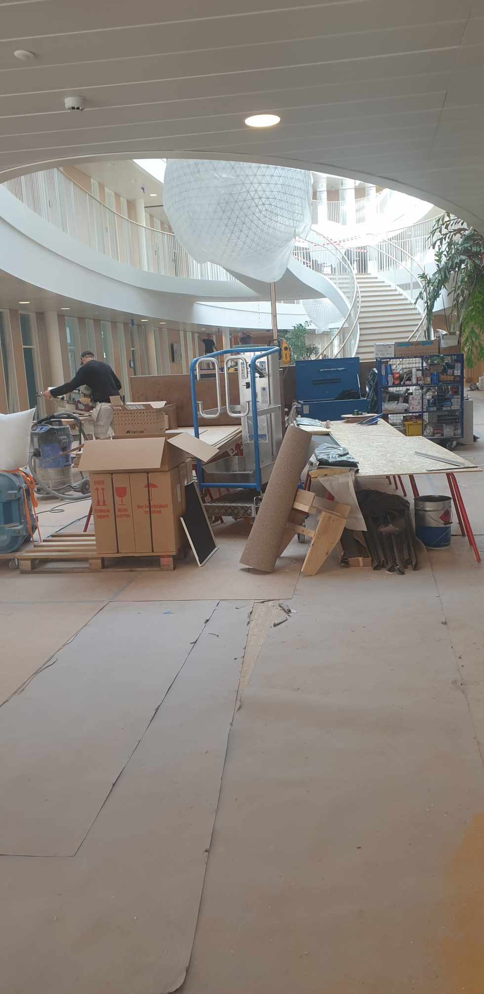 Wnętrze budynku w trakcie prac wykończeniowych: widoczny pracownik szlifujący ścianę, podnośnik, kartony, narzędzia i materiały budowlane na podłodze, w tle spiralne schody.